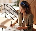 Woman in blazer writing notes while using smartphone in sunlit cafe.