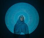 A woman in a hijab is illuminated by a circular Arabic calligraphy projection.